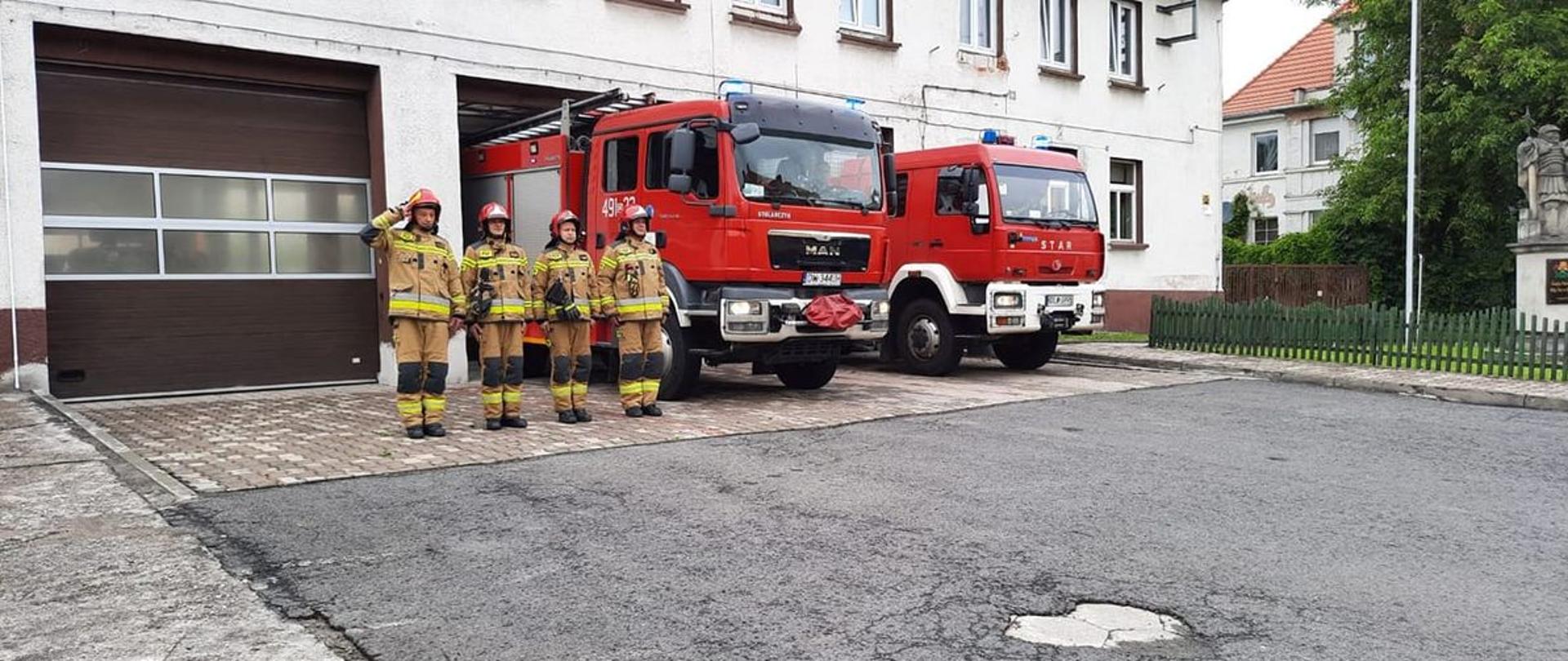 Zdjęcie przedstawia strażaków z Jednostki Ratowniczo-Gaśniczej Państwowej Straży Pożarnej w Lwówku Ślaskim podczas uczczenia 77 rocznicy Powstania Warszawskiego. Strażacy stoją w szeregu na placu zewnętrznym jednostki. W tle widoczne znajdują się samochody pożarnicze. 