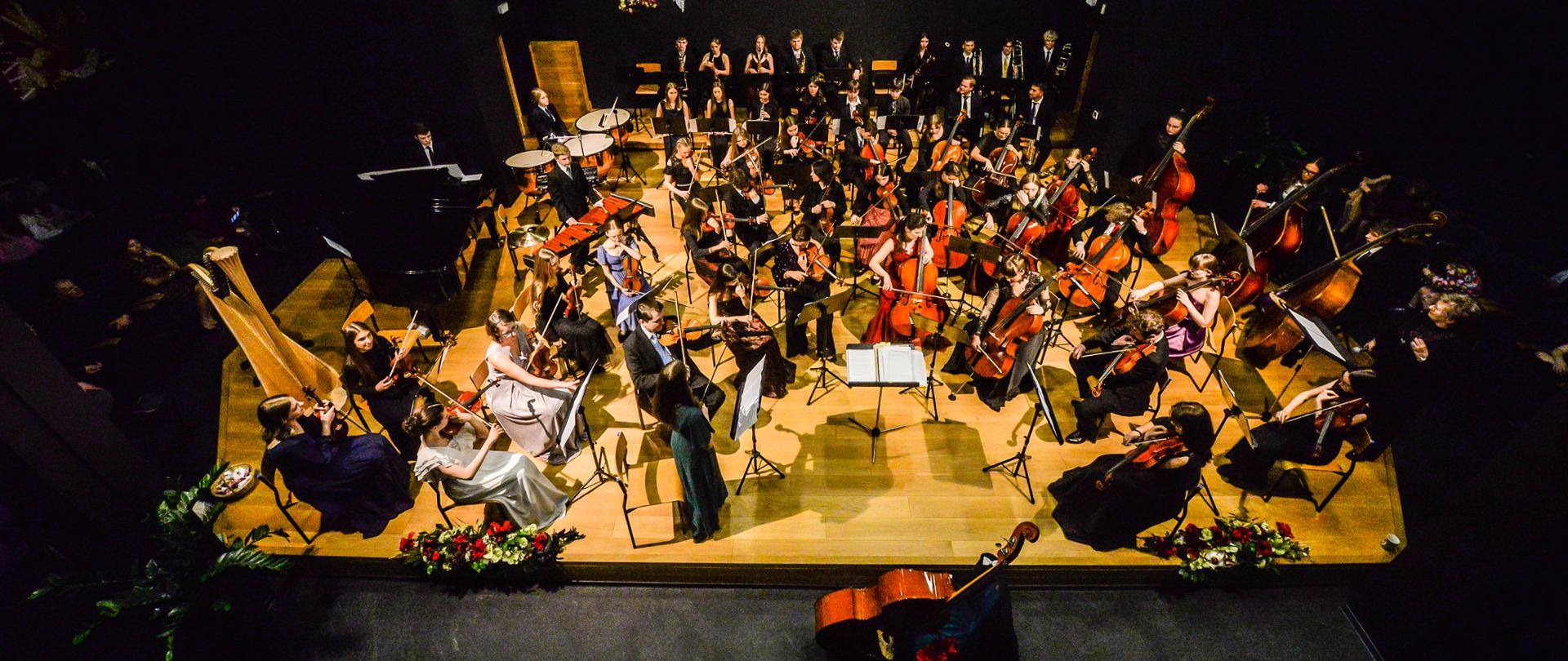 Kolorowe zdjęcie Orkiestry Symfonicznej Szkoły Szafranków na scenie w sali koncertowej zrobione z lotu ptaka. Wszyscy członkowie orkiestry ubrani są w odświętne stroje (chłopcy w ciemne garnitury, białe koszule i kolorowe krawaty, dziewczyny w kolorowe balowe suknie). Scena udekorowana jest kwiatami. Przed sceną znajduje się tematyczna dekoracja związana z ostatkami tj. Kontrabas oparty o przystrojone krzesło w zielone płótno i kwiaty.