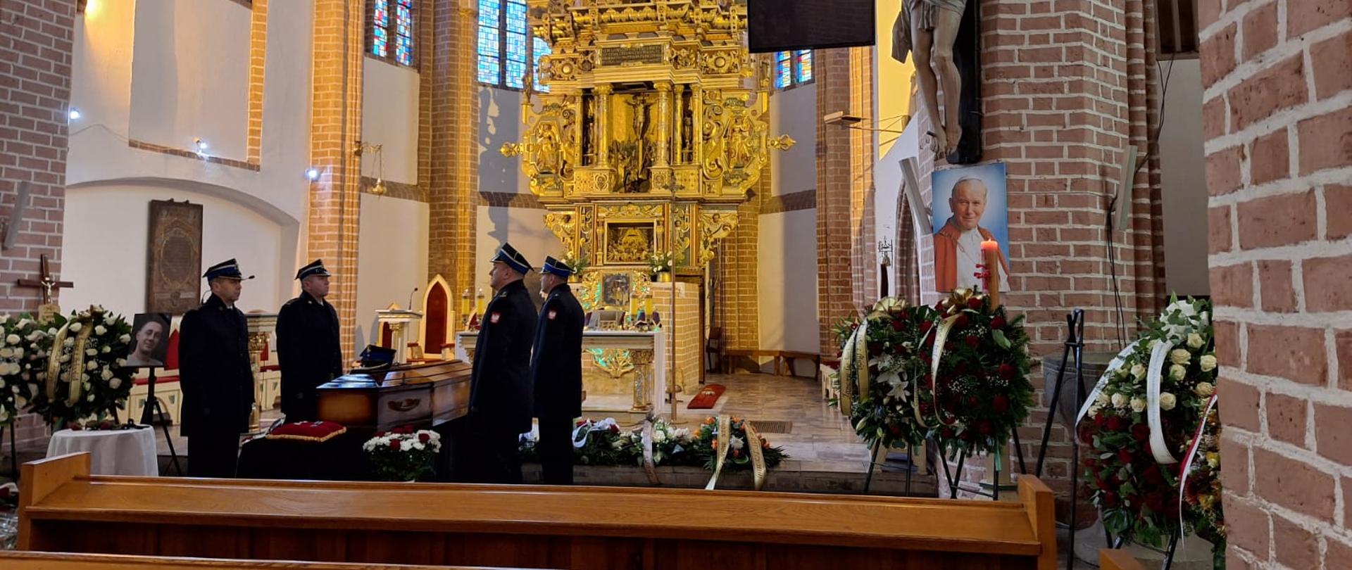 Zdjęcie przedstawia wnętrze kościoła o wysokich, ceglastych filarach i kolorowych witrażach. W centralnej części widać bogato zdobiony, złoty ołtarz sięgający wysokości kilku metrów. Przed ołtarzem stoi trumna przykryta flagą, a obok niej czterech funkcjonariuszy w galowych, ciemnych mundurach pełni wartę honorową. Po prawej stronie, przy jednym z filarów, znajduje się duża figura Jezusa ukrzyżowanego. Pod nią ustawiono portret uśmiechniętego, starszego duchownego w czerwonej sutannie oraz liczne wieńce z kwiatów. Na pierwszym planie znajdują się drewniane ławki kościelne. Cała scena ma uroczysty, podniosły i pogrzebowy charakter.