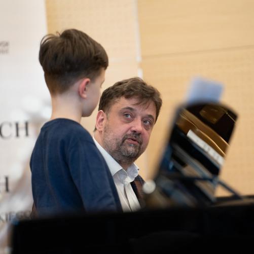 zdjęcie przedstawia prowadzącego warsztaty w rozmowie z uczestnikiem warsztatów przy dwóch fortepianach na scenie sali koncertowej, ujęcie z bliska