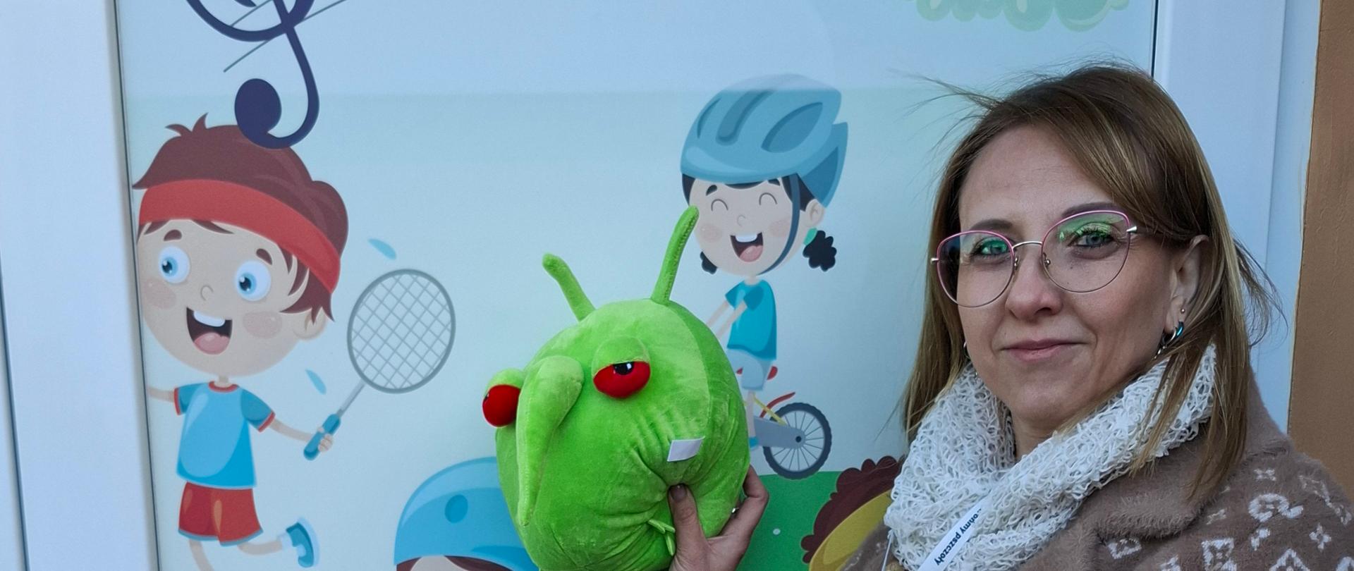 Inspektor stojąca na tle kolorowych do przedszkola i trzymająca w ręce maskotkę PESTY 