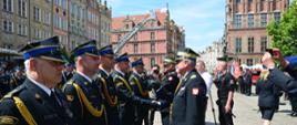 Strażacy w mundurach galowych stoją obok siebie przed nimi kadra kierownicza Państwowej Straży Pożarnej wręcza odznaczenia oraz gratuluje obok stoją przedstawiciele mediów.
