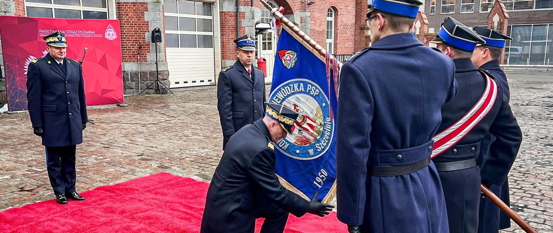 Jeden ze strażaków klęczy na czerwonym dywanie przed sztandarem. Trzyma go dłoniami. 