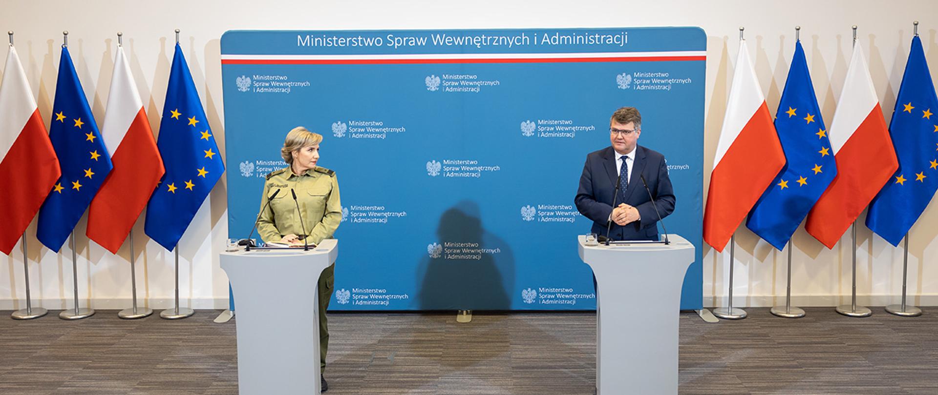 Na zdjęciu widać wiceministra Macieja Wąsika i gen. bryg. SG Wioletę Gorzkowską stojących za mównicami w trakcie konferencji prasowej. W tle widać ściankę MSWiA, flagi Polski i UE .