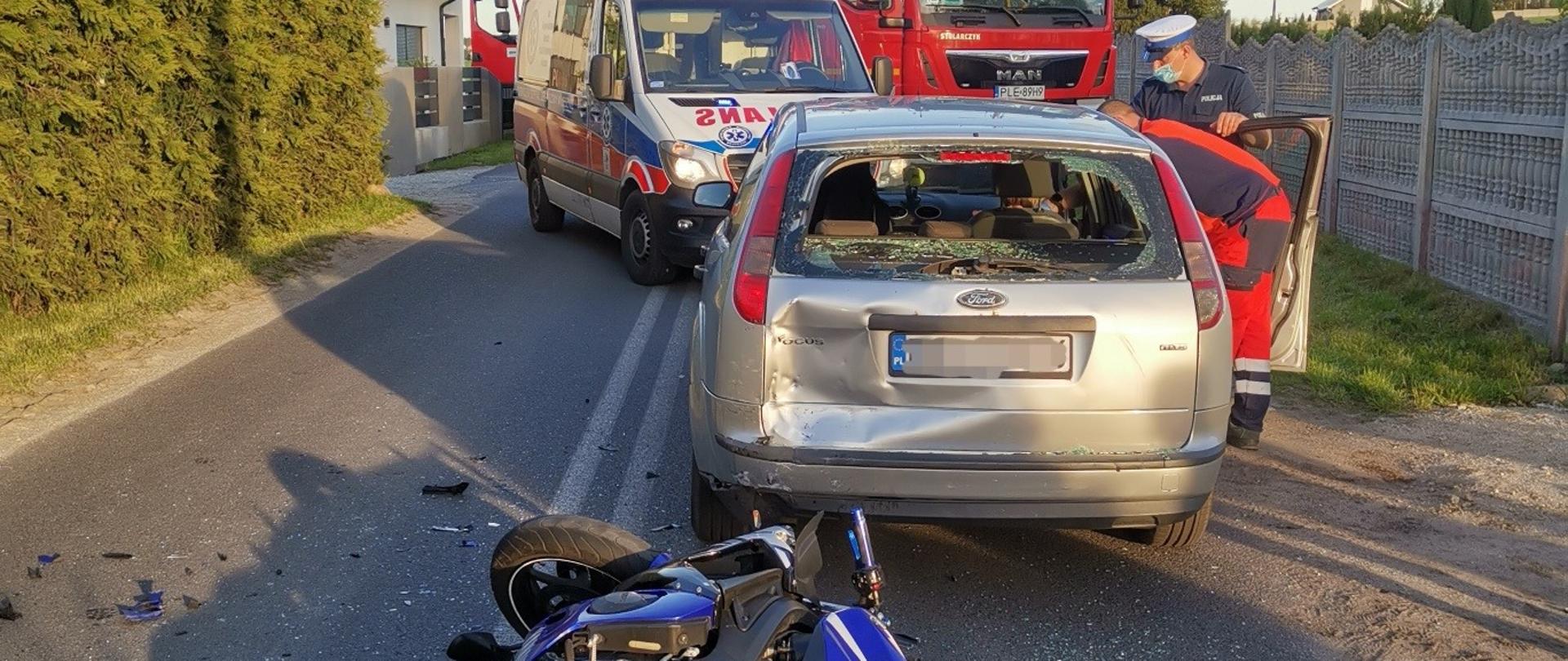 Motocykl leży na drodze. Obok stoi samochód osobowy z uszkodzoną szybą tylną. W tle widoczna karetka oraz samochód straży pożarnej.