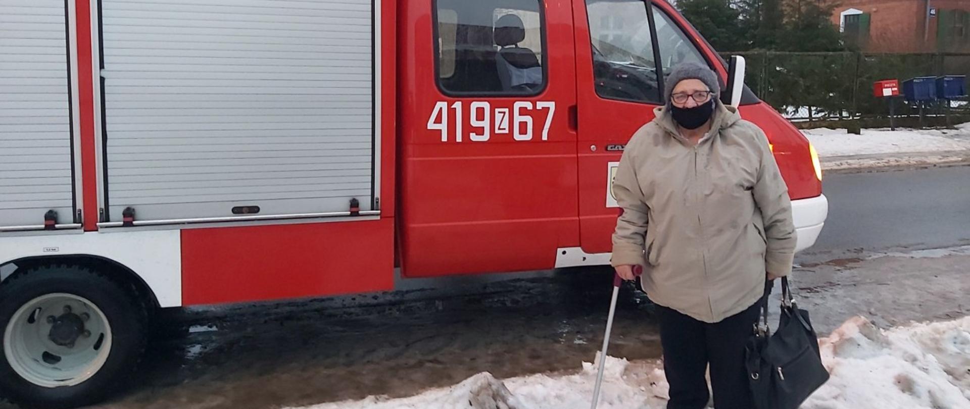 Na zdjęciu znajduje się starsza kobieta, ubrana w beżową kurtkę, trzymająca w lewej ręce czarną torebkę, a w prawej trzyma kulę. Ma ubraną na twarzy maseczkę. Za kobietą stoi wóz strażacki, a za pojazdem znajdują się domy. Dookoła jest śnieg.