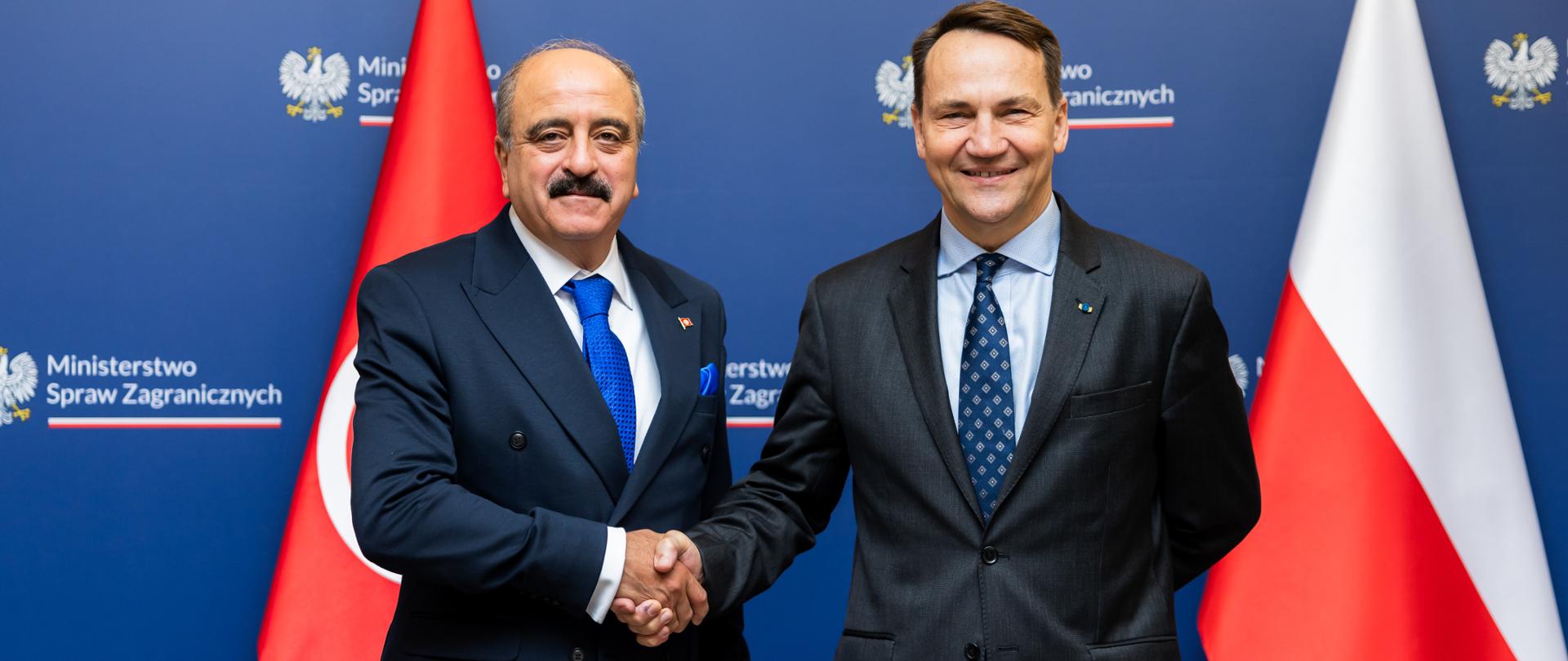 Na zdjęciu szefowie dyplomacji Polski i Tunezji podczas uścisku dłoni. W tle ścianka z logotypami MSZ i dwie flagi: Polski i Tunezji.