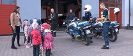 dzieci z opiekunami stoją na zewnątrz przed bramą garażową i oglądają dwa nowe motory należące do powiatowej komendy policji dwaj policjanci opowiadają o nich dzieciom