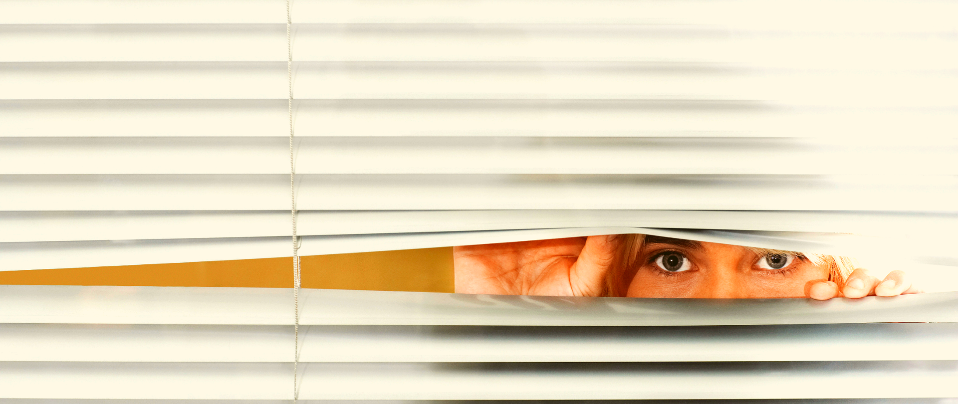 Kobieta spoglądająca pomiędzy żaluzjami. Widoczny fragment dłoni przytrzymujących żaluzje, oczy i fragment nosa.