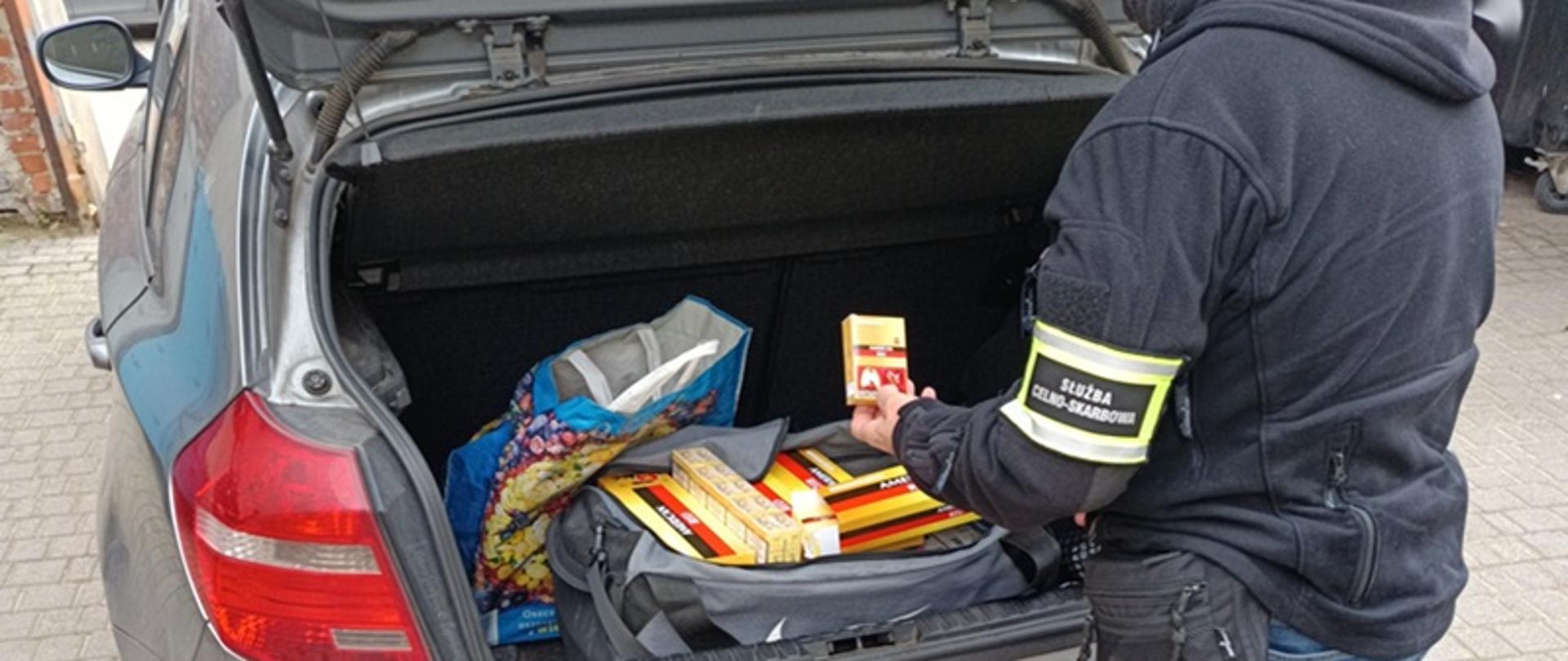 Funkcjonariusz ogląda paczki papierosów umieszczone w otwartym bagażniku auta osobowego. 