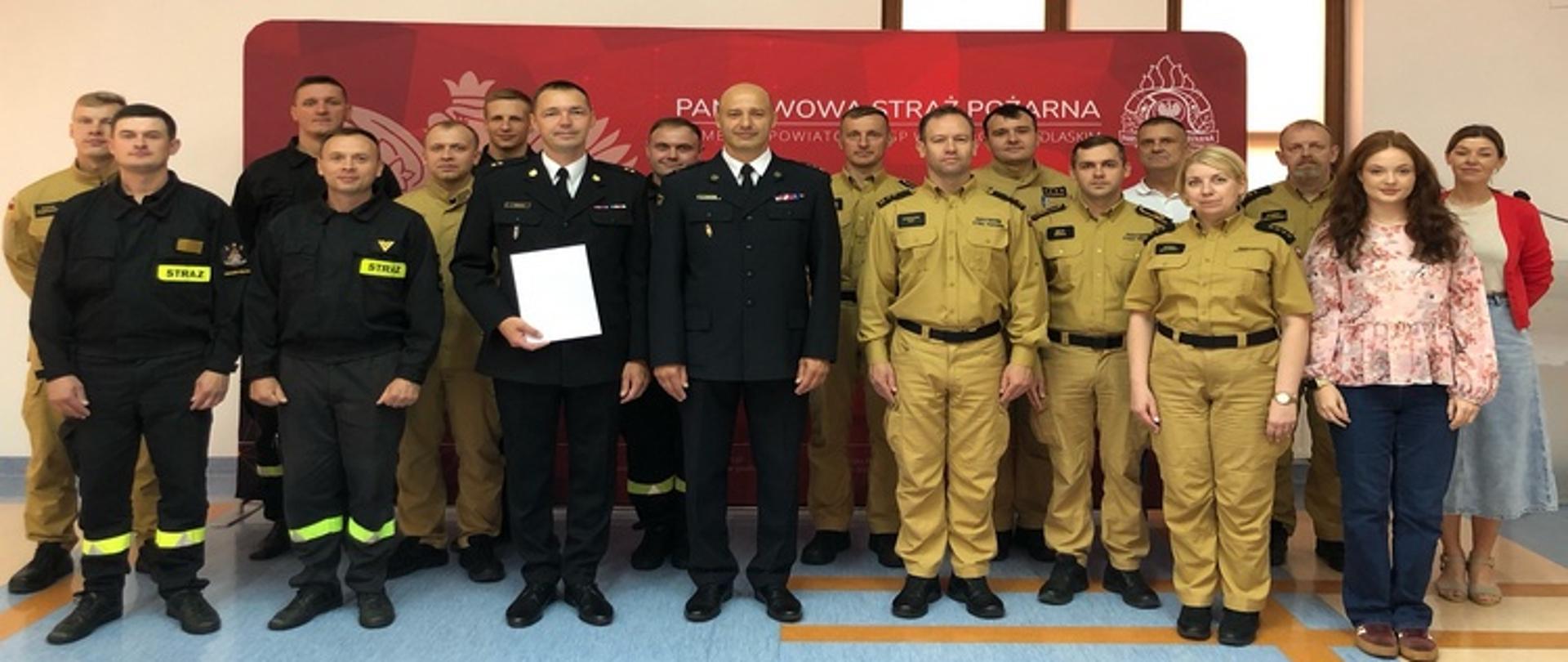 Zdjęcia przedstawia funkcjonariuszy PSP w umundurowaniu służbowym koloru piaskowego oraz w mundurach galowych koloru granatowego. Stoją na tle czerwonego bilbordu z logiem Państwowej Straży Pożarnej. Zdjęcie wykonane w sali szkoleniowej, ściany koloru beżowego.