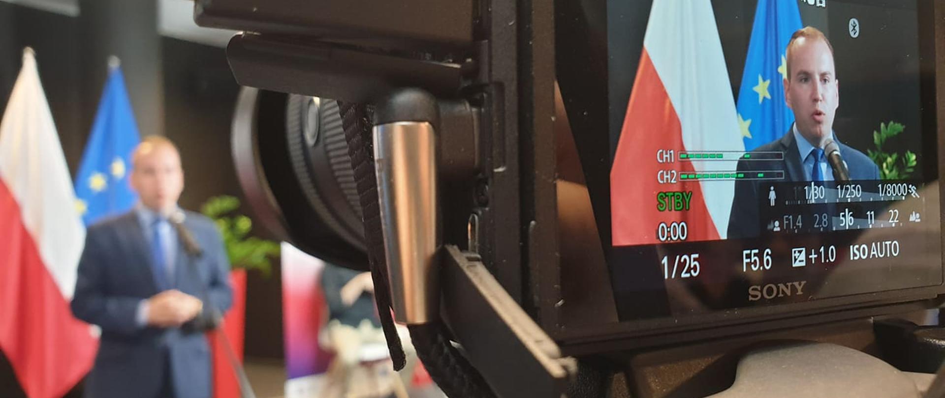 Konferencja prasowa ministra Adama Andruszkiewicza. Widok na aparat fotograficzny w tle flagi, przed nimi stoi minister Andruszkiewicz.