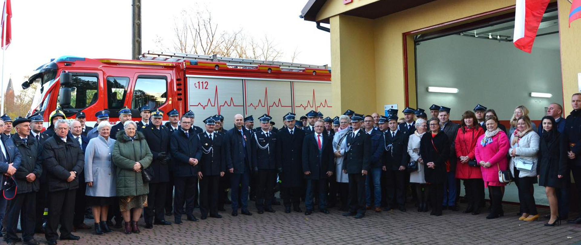 Zdjęcie grupowe uczestników uroczystości 75 lecia OSP Kowalewice. W tle remiza i samochód ratowniczo gaśniczy