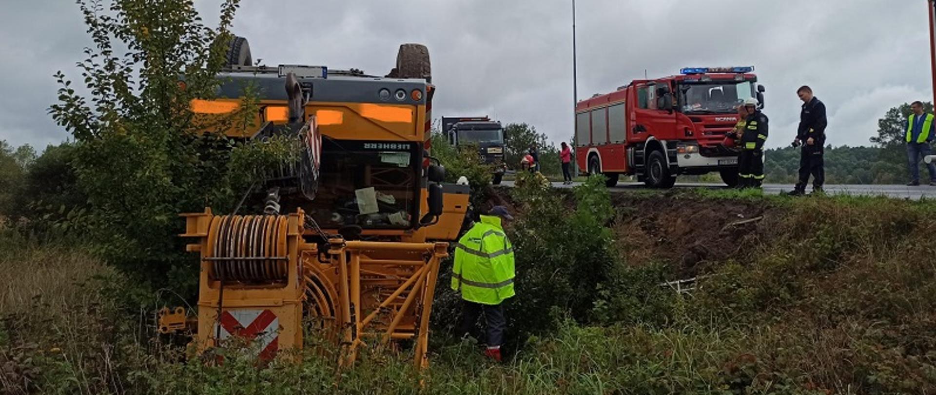 Zdjęcie przedstawia widoczny od przodu żółty dźwig budowlany na samochodzie ciężarowym leżący do góry kołami na poboczu drogi. Po prawej stronie pojazdu strażacy i policjanci na tle czerwonego samochodu strażackiego