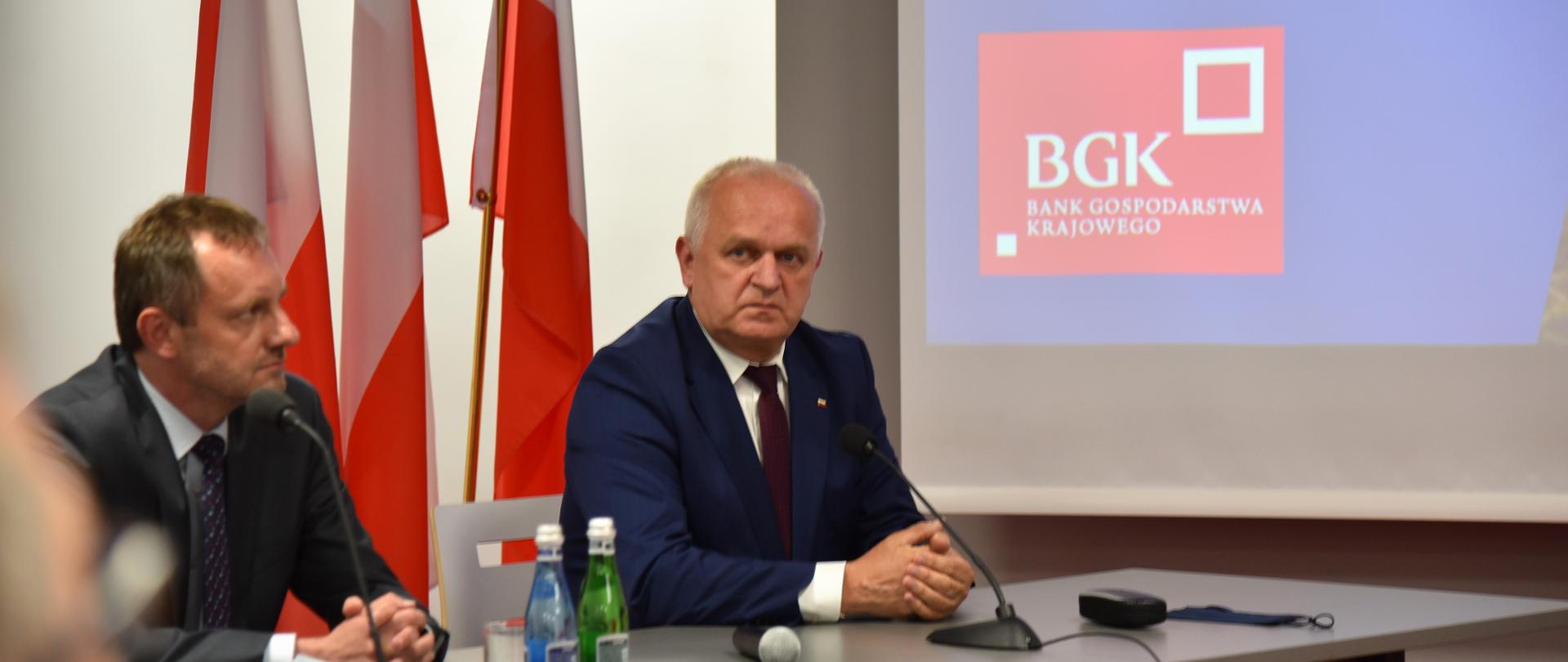 Wojewoda z Prezesem BGK siedzą przy stole konferencyjnym. W tle biało-czerwone flagi i fragment prezentacji z czerwono-białym logiem BZK