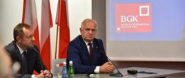 Wojewoda z Prezesem BGK siedzą przy stole konferencyjnym. W tle biało-czerwone flagi i fragment prezentacji z czerwono-białym logiem BZK