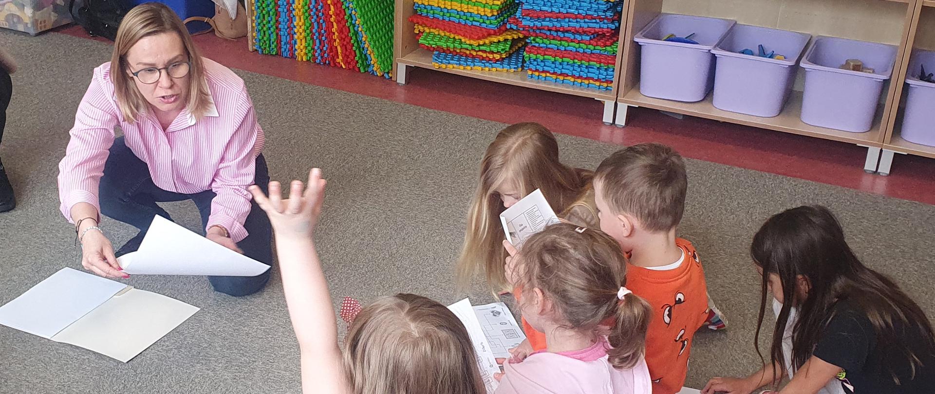 Gromadka dzieci siedząca na podłodze w sali przedszkolnej, obok dwie panie inspektor. Jedna z dziewczynek podnosi rękę do góry.