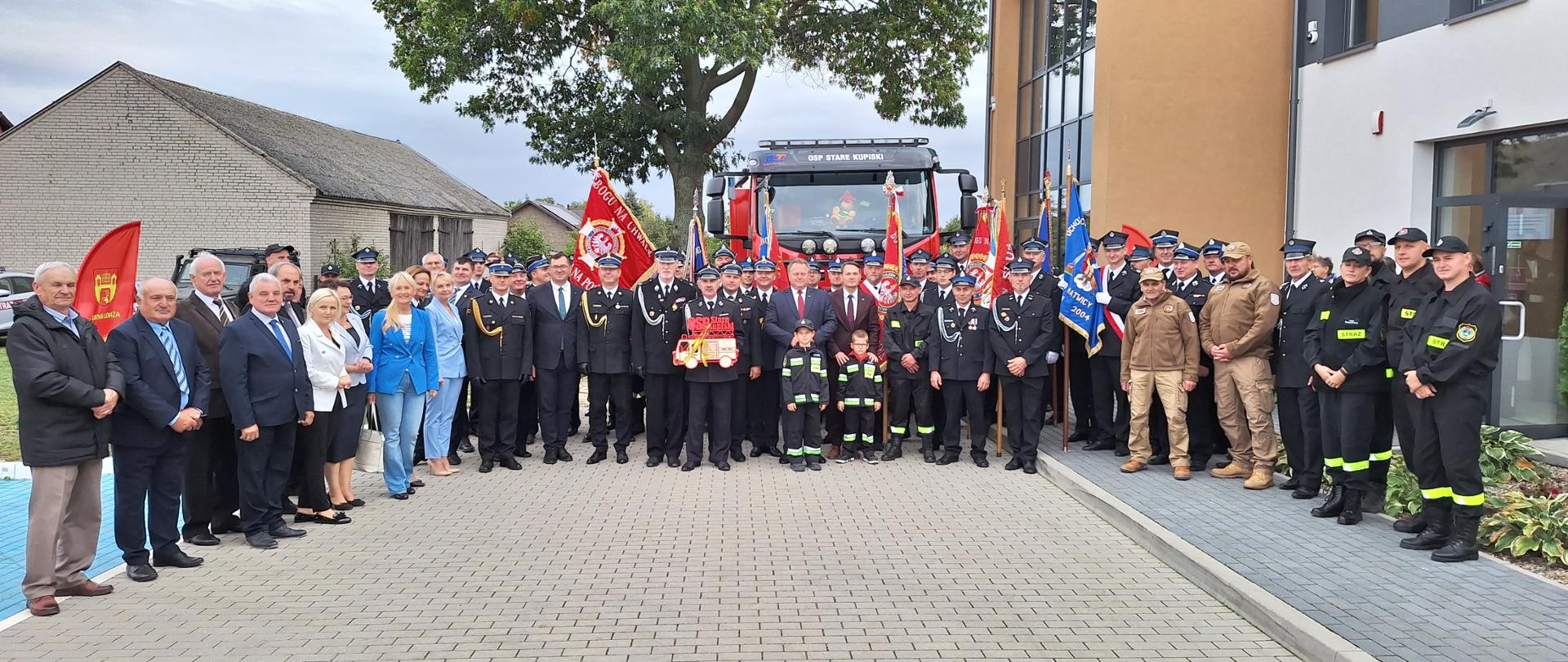 Zdjęcie grupowe uczestników uroczystości na placu przed budynkiem remizy Ochotniczej Straży Pożarnej w Starych Kupiskach. W tle widoczny przekazany samochód ratowniczo-gaśniczy