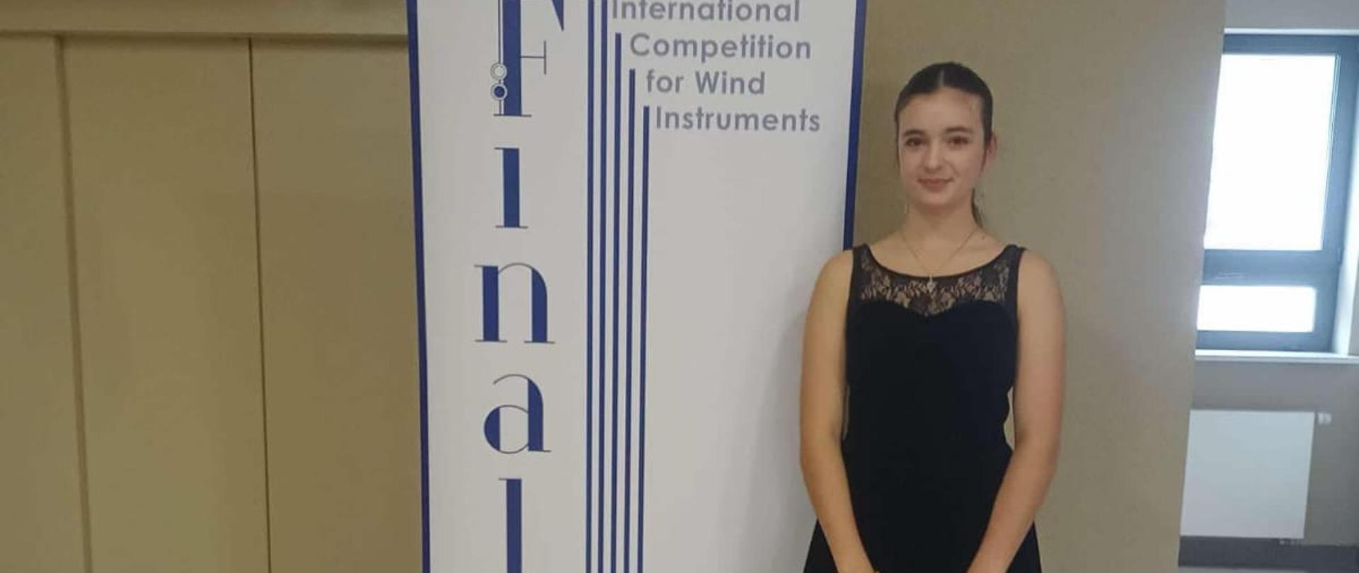 Zosia Zając stojąca z fletem w futerale na korytarzu przy plakacie konkursu Finalis.
