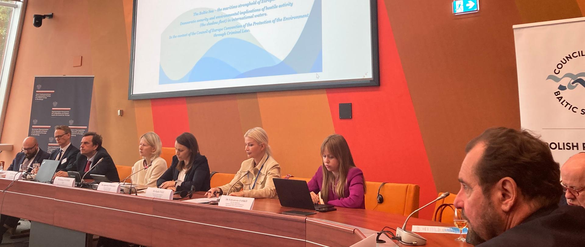 Stół przy którym obradują uczestnicy debaty ""„Bałtyk – europejska twierdza Europy. Bezpieczeństwo demokratyczne i konsekwencje dla środowiska spowodowane aktywnością floty cieni na wodach międzynarodowych. W kontekście Konwencji o Ochronie Środowiska przez Prawo Karne”.