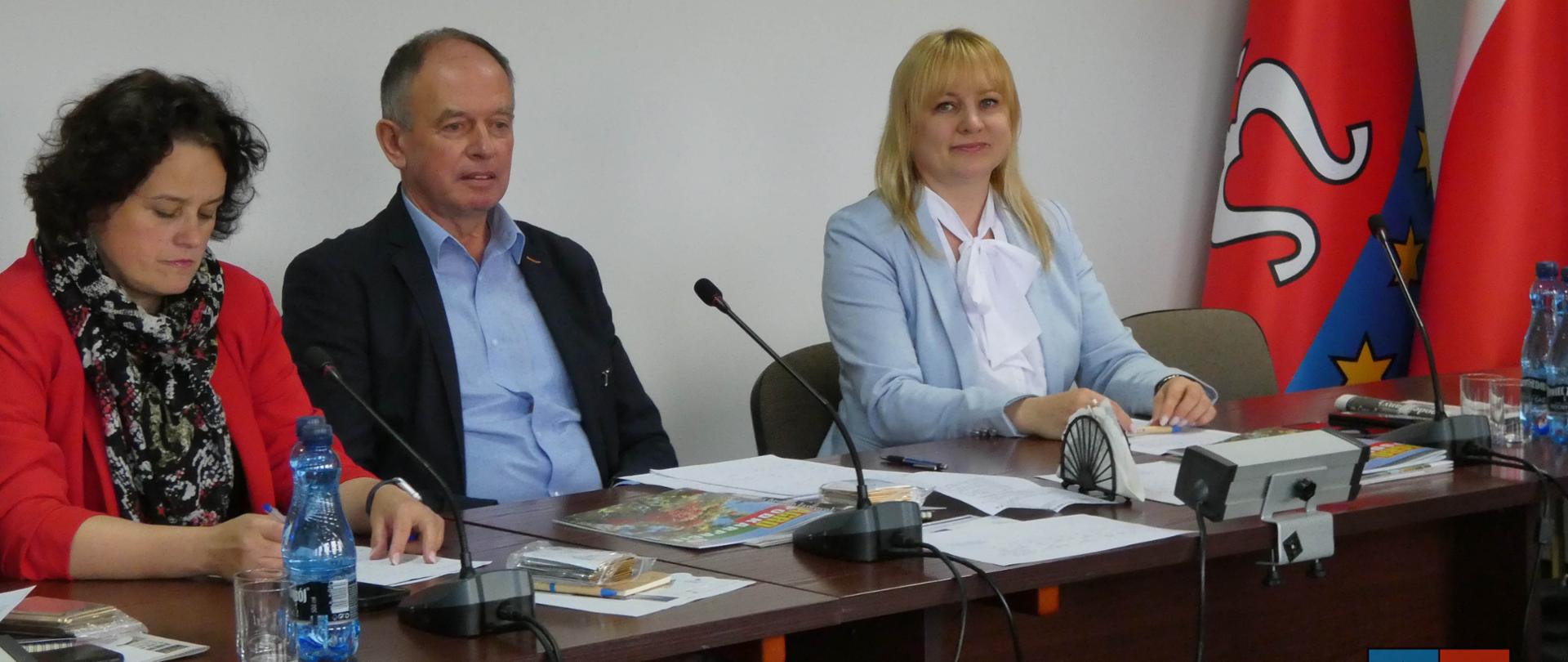 Trzy osoby siedzące w jasnym pomieszczeniu. Dwie kobiety i jeden mężczyzna. Kobieta po lewej stronie ubrana w czarną bluzkę i czerwoną marynarkę. Kobieta po prawej ubrana w jasno niebieski garnitur i białą koszulę. Mężczyzna w środku ubrany w czarna marynarkę i niebieską koszulę. Siedzą przed stołem na którym lezą dokumenty. 