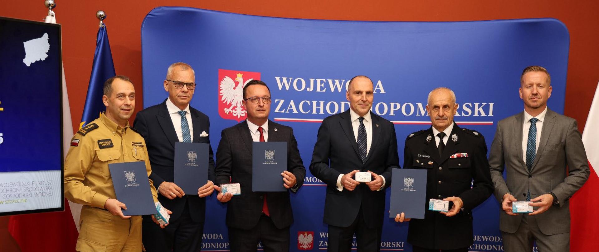 Na zdjęciu widzimy sześciu mężczyzn stojących naprzeciw aparatu. Są oni na tle niebieskiego banera z napisem "Wojewoda Zachodniopomorski”. 