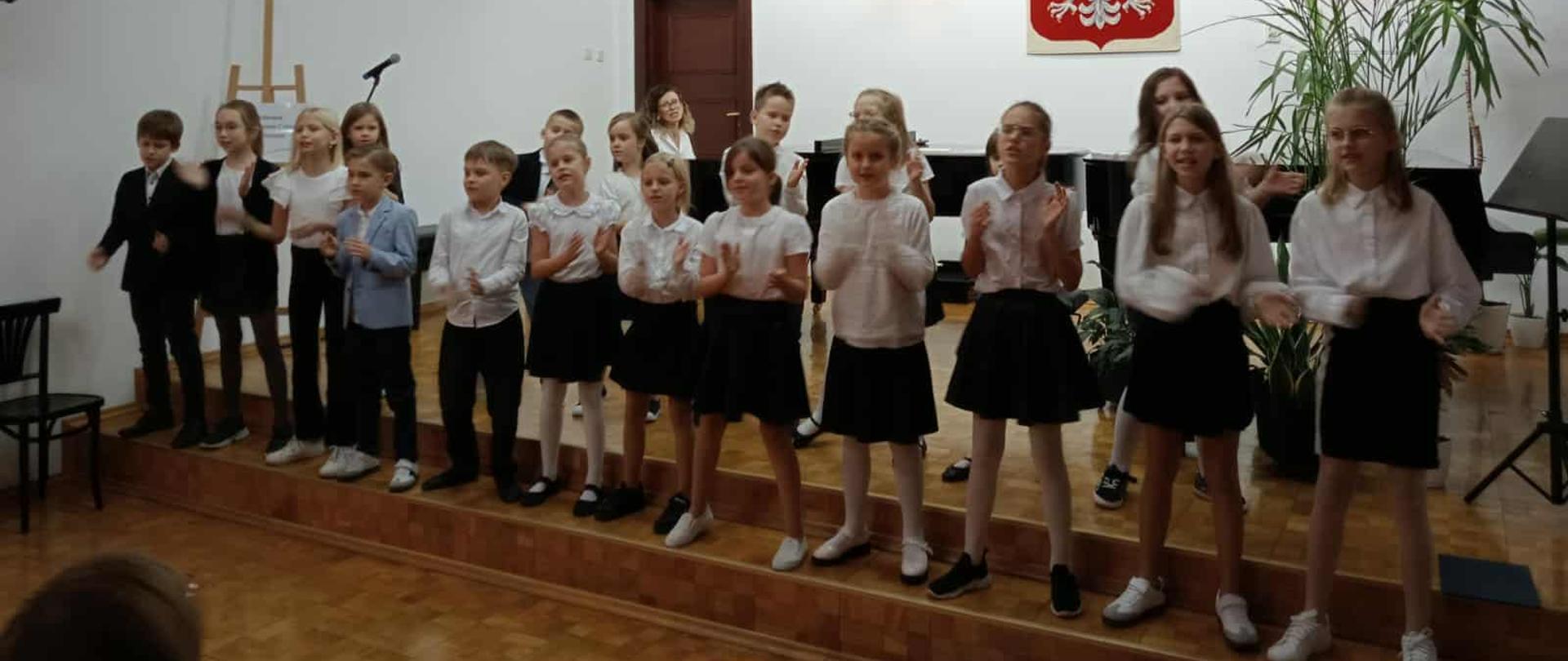 Pierwszoklasiści stoją na scenie w sali koncertowej i śpiewają. Są odświętnie ubrani w białe koszule, ciemne spódnice i spodnie. W tle widoczne jest godło Polski i zielone rośliny w doniczce.