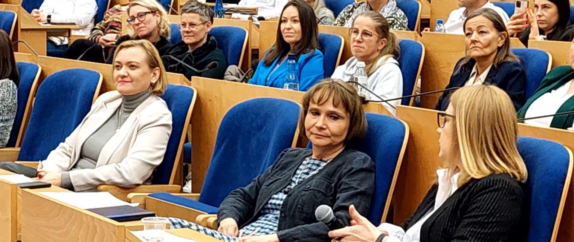 wiele osób siedzi w dużej sali i słucha wypowiedzi prelegenta