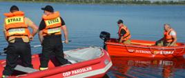 Ćwiczenia z udziałem SGRWN. Zdjęcie przedstawia zbiornik retencyjny w Jutrosinie. Na nim zwodowane są dwie jednostki pływające koloru czerwonego: ponton z białym napisem "OSP Pakosław" i łódka z napisem "OSP Jutrosin Straż". Na pokładzie pontonu stoi dwóch, a w łodzi siedzi dwóch strażaków. Wszyscy mają założone kapoki koloru pomarańczowego z napisem "STRAŻ". W oddali widać drzewa.