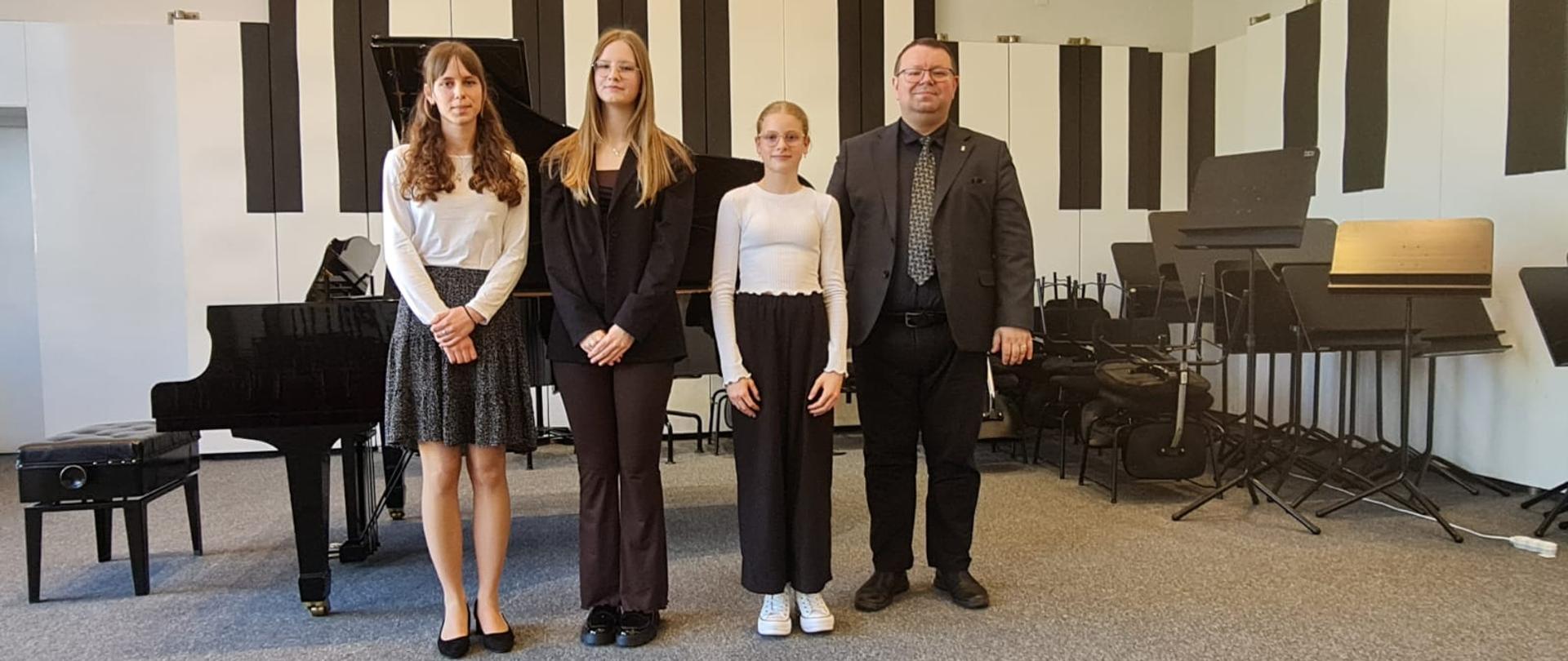 Mężczyzna i trzy nastolatki stoją na scenie przed fortepianem.