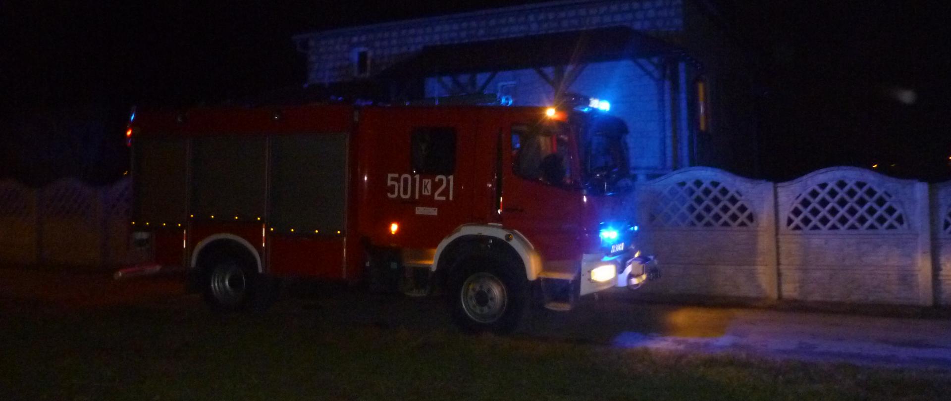 Samochód pożarniczy z włączonymi sygnałami świetlnymi koloru niebieskiego w tle ogrodzenie betonowe oraz dom