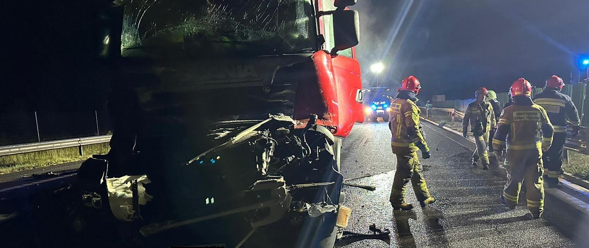 Zdjęcie strażaków interweniujących przy kolizji dwóch ciężarówek. Jedna z nich ma mocno uszkodzony przód pojazdu, a strażacy poruszają się wokół niego. W tle widać samochód strażacki. 