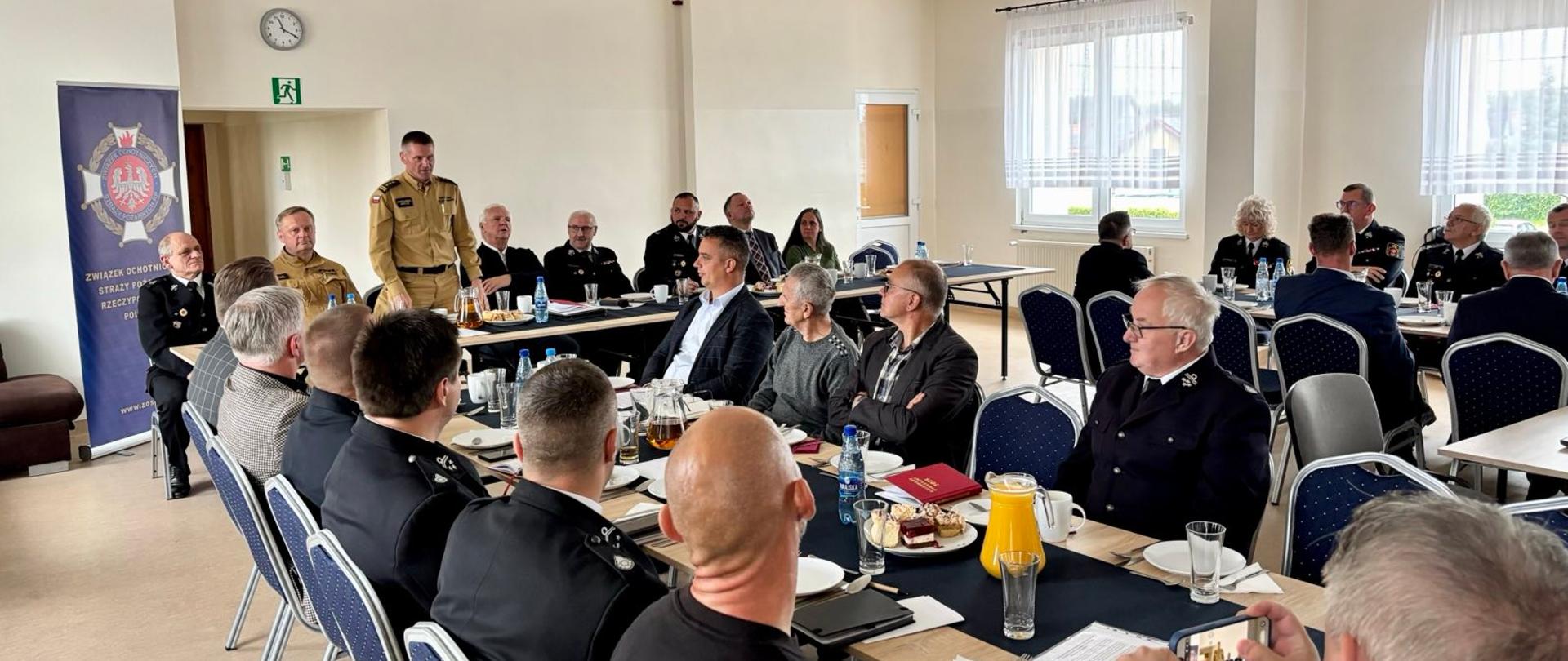 Widok na salę obrad, stoły ustawione w rzędach przy stole prezydialnym, uczestnicy posiedzenia w mundurach, po lewej stronie sali baner ZOSP