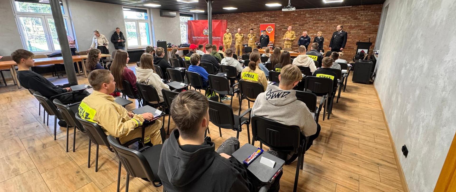 Uczestnicy turnieju siedzą w sali szkoleniowej. Z przodu stoi komisja oraz komendant.