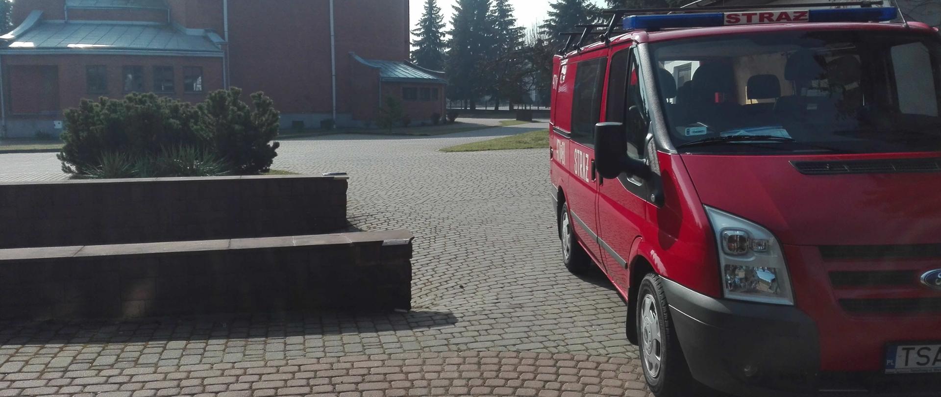Zdjęcie przedstawia samochód strażacki, który stoi na placu obok kościoła. W tle widać murowany kościół.