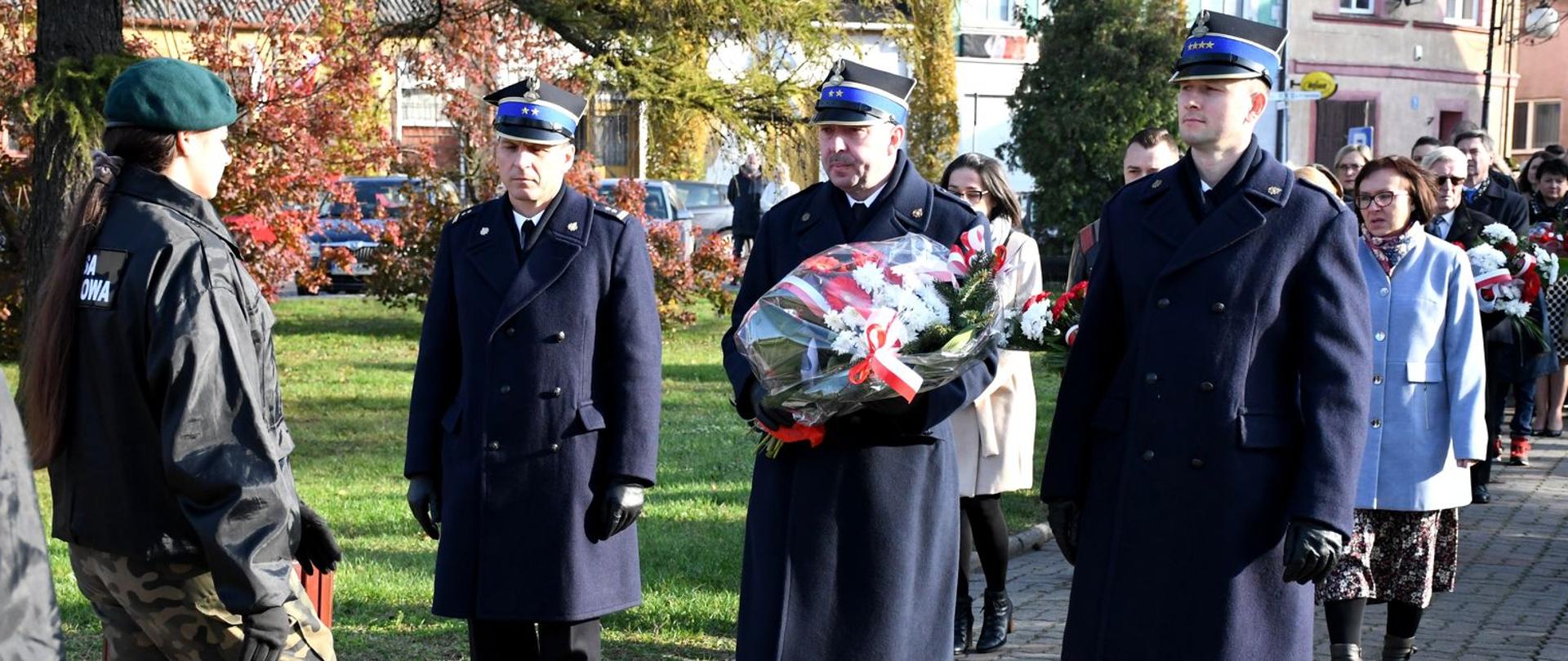 Na zdjęciu trzej strażacy ubrani w płaszcze zimowe, na głowach czapki rogatywki, stoją przed pomnikiem. Jeden ze strażaków trzyma wiązankę kwiatów.