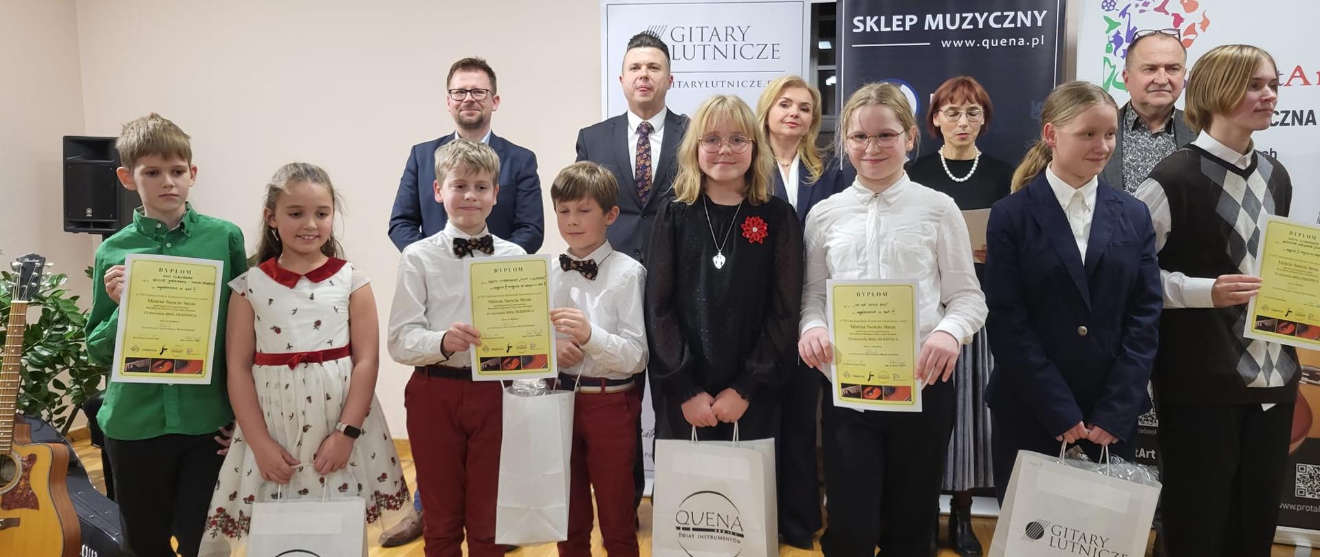 Grupa uczniów wraz jurorami konkursowymi pozują do zdjęcia grupowego trzymając nagrody oraz dyplomy za udział w Ogólnopolskim Konkursie Gitarowym o tytuł "Mistrza Sześciu Strun" w Oleśnicy - 13 stycznia 2024