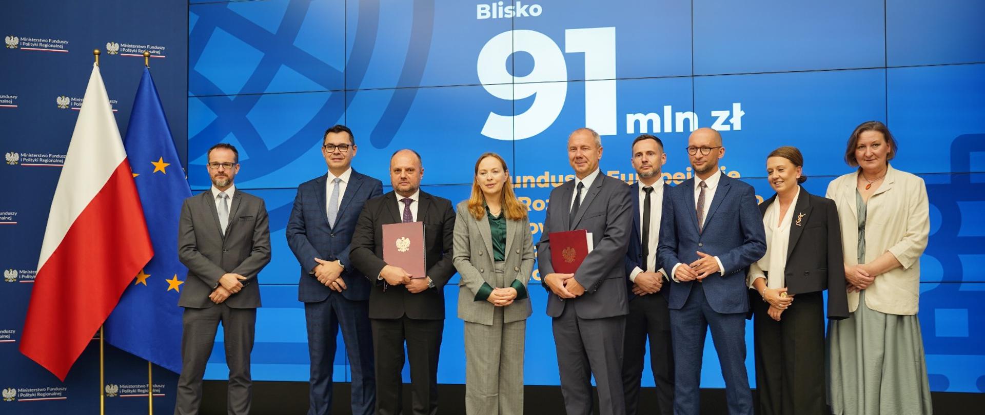 Dziewięć osób stoi obok siebie na scenie. Czwarta od lewej stoi lekko uśmiechnięta minister Katarzyna Pełczyńska-Nałęcz.