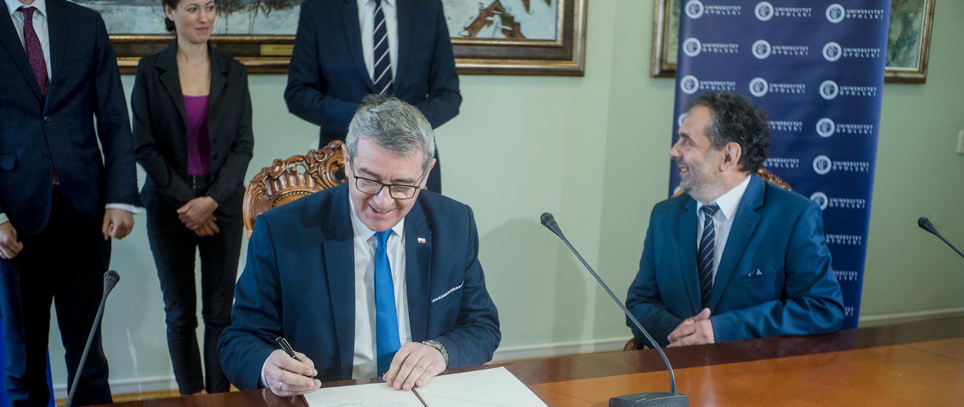 Minister siedzi za stołem, przed nim książka i mikrofon.