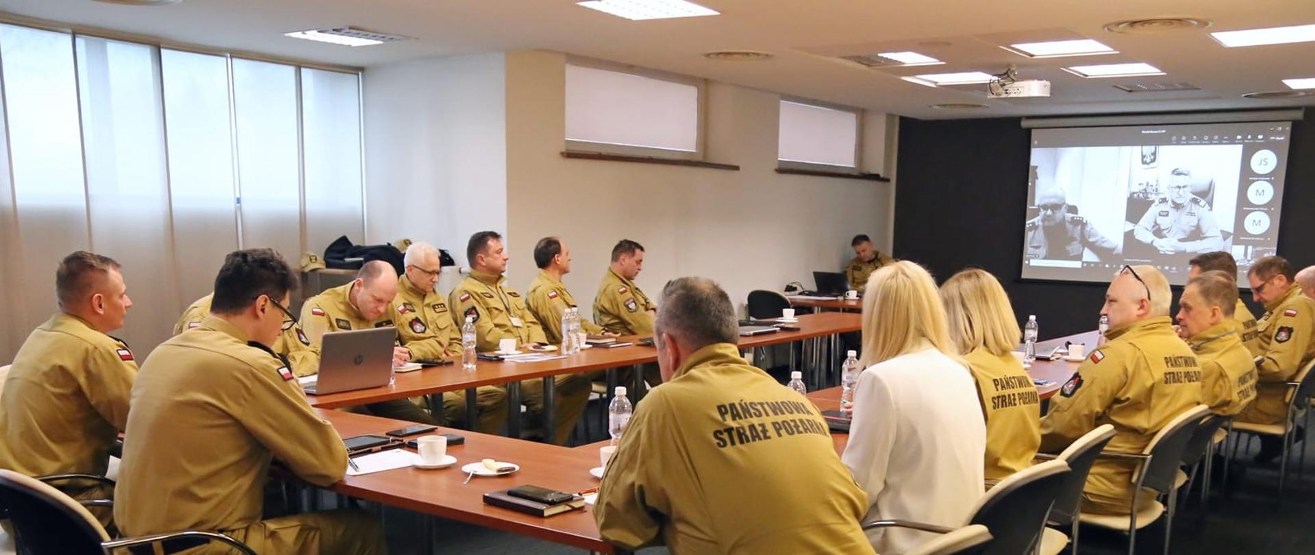 W sali przy stole w kształcie litery U siedzą funkcjonariusze Szkoły , uczestniczący w naradzie rocznej. Z przodu sali na ekranie widać zastępcę komendanta głównego PSP oraz dyrektora biura edukacji KG PSP, którzy uczestniczą w naradzie w formie wiedokonferencji