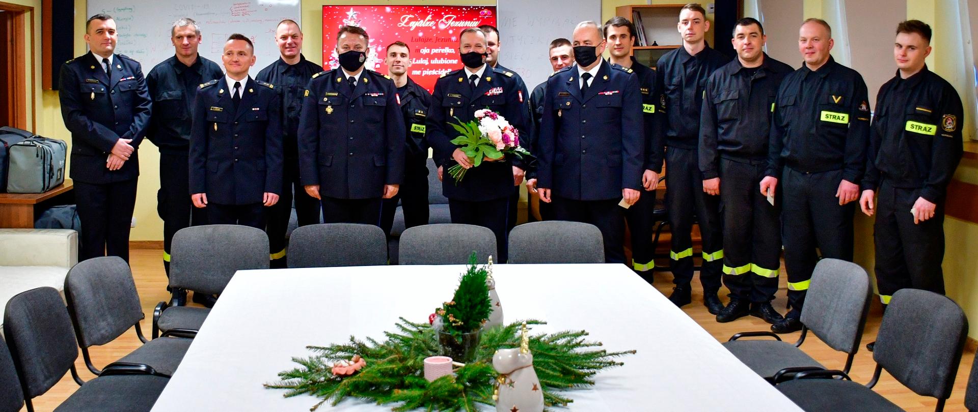 Na zdjęciu widać grupę 15 strażaków, którzy stoją za świątecznym stołem nakrytym białym obrusem. W środku zdjęcia widać świąteczny stroik.