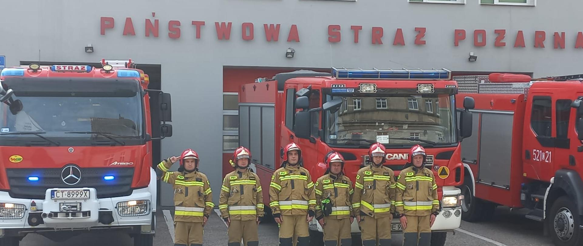 Zdjęcie wykonano w ciągu dnia przed budynkiem Komendy Powiatowej Państwowej Straży Pożarnej w Sępólnie Krajeńskim. Strażacy Państwowej Straży Pożarnej ubrani w ubrania specjalne i hełmy stoją na baczność w szeregu przed samochodami ratowniczo- gaśniczymi stojącymi przed garażami. 