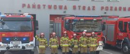 Zdjęcie wykonano w ciągu dnia przed budynkiem Komendy Powiatowej Państwowej Straży Pożarnej w Sępólnie Krajeńskim. Strażacy Państwowej Straży Pożarnej ubrani w ubrania specjalne i hełmy stoją na baczność w szeregu przed samochodami ratowniczo- gaśniczymi stojącymi przed garażami. 