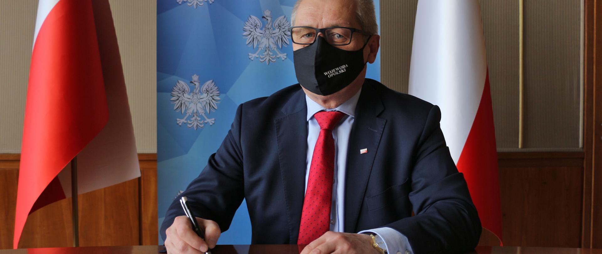 Wojewoda opolski podpisuje decyzje o uruchomieniu kolejnych Punktów Szczepień Powszechnych. Wojewoda siedzi przy półokrągłym stole, w dłoni ma pióro. Przed nim na stole czerwona teczka z dokumentem. Wojewoda spogląda w obiektyw, na nosie ma okulary i maseczkę. W tle roll up z napisem WOjewoda OPolski oraz biało-czerwona flaga.