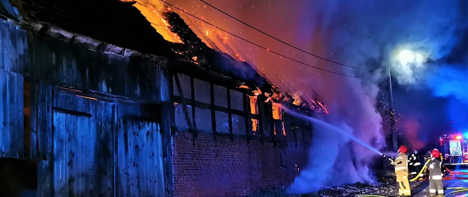 Zdjęcie przedstawia palącą się stodołę i podających jeden prąd wody strażaków.