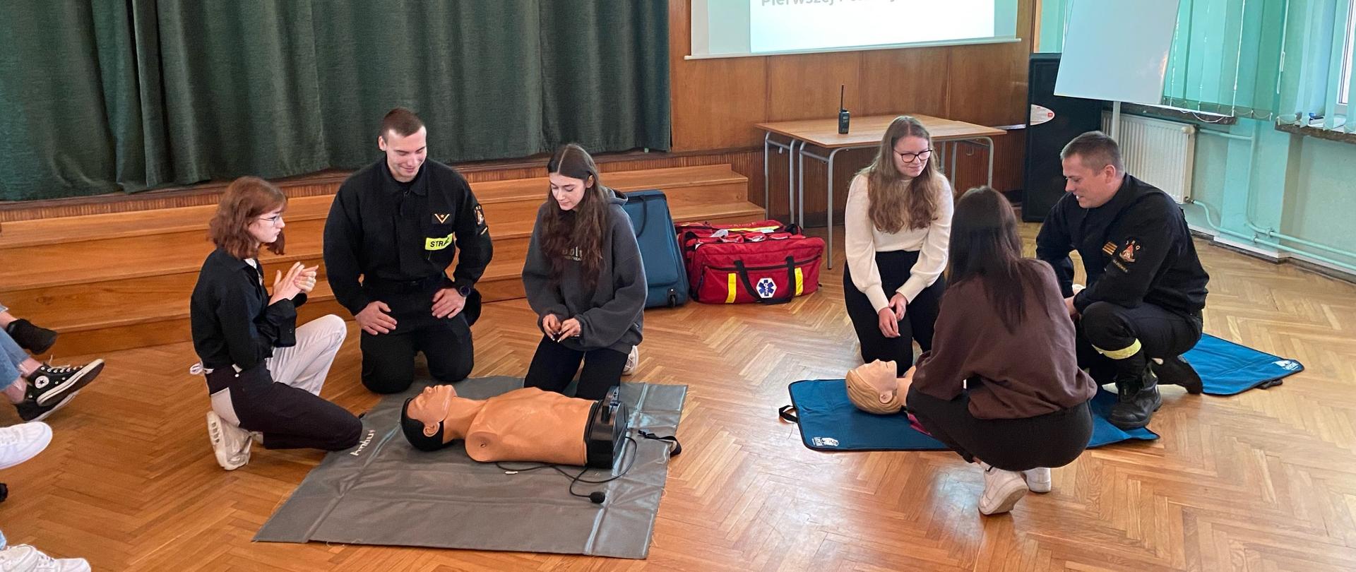 W pomieszczeniu przy dwóch fantomach klęczą po trzy osoby. Wśród każdej z nich znajduje się strażak w czarnym ubraniu.