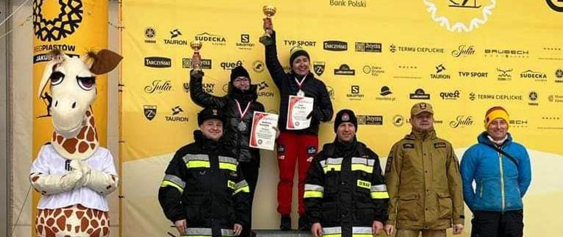 Strażacy w mundurach oraz w strojach sportowych stoją przy/na podium