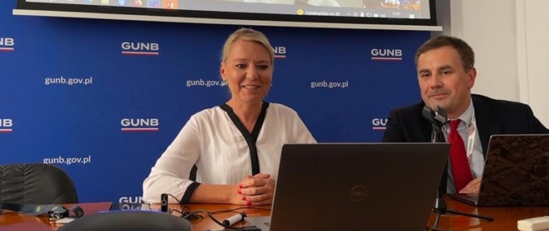 Dwóch prelegentów siedzących przy stole z laptopami i mikrofonami, na tle niebieskiej ścianki z logo „GUNB” i adresem „gunb.gov.pl”; nad nimi ekran z prezentacją.
