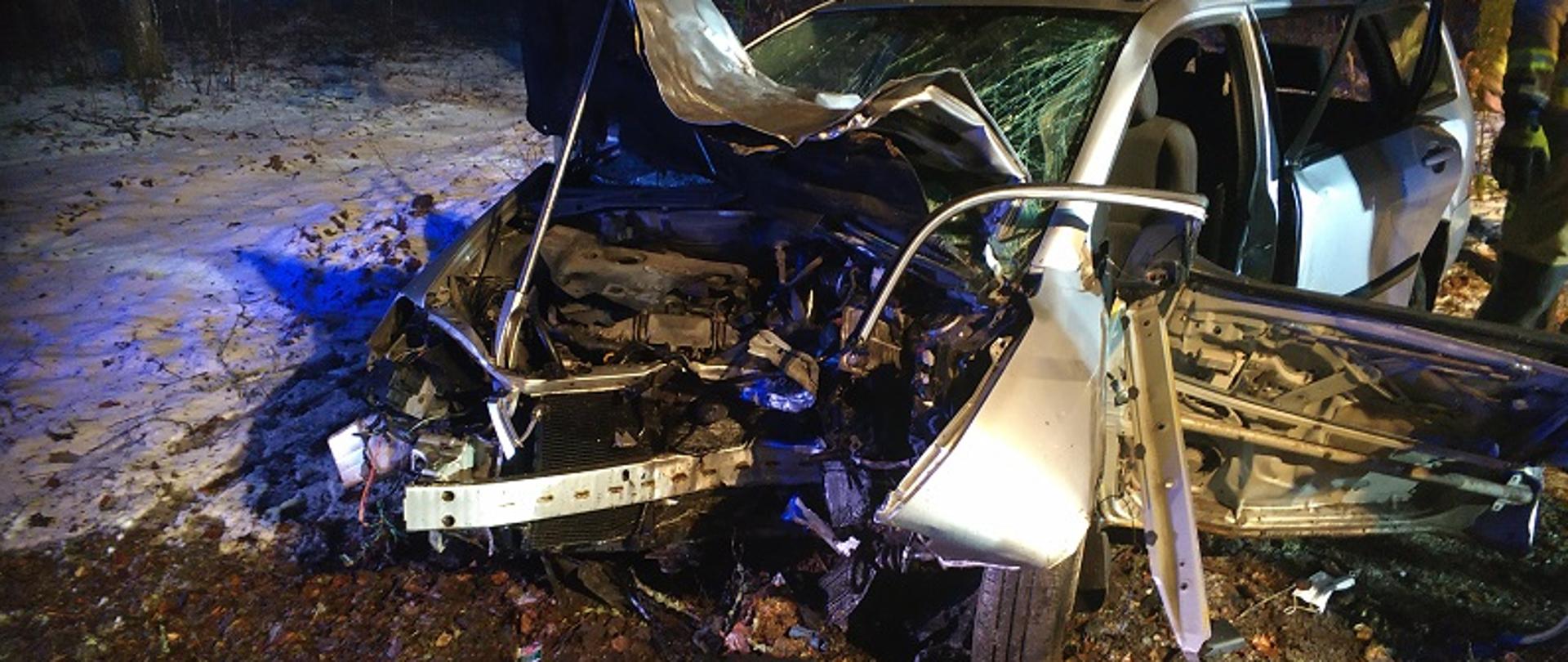 Zdjęcie wykonane nocą, na pierwszym planie widoczny przodem wrak samochodu osobowego Toyota Corolla koloru srebrnego. Na tle drzew lasu widnieje otwarta maska przednia komory silnikowej podparta łomem strażackim typu „Hooligan”, rozbita szyba przednia i otwarte drzwi pojazdu od strony kierowcy