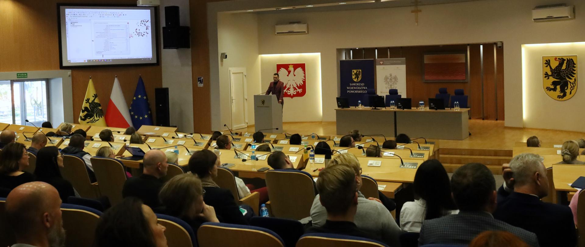 Sala kinowa w brązowo-białych odcieniach. Na pierwszym planie widać ludzi siedzących na krzesłach. W oddali za mównicą stoi mężczyzna na scenie. Po prawej stronie stoi stół prezydialny. Po lewej stronie na ścianie wisi ekran, na którym wyświetlana jest prezentacja. 